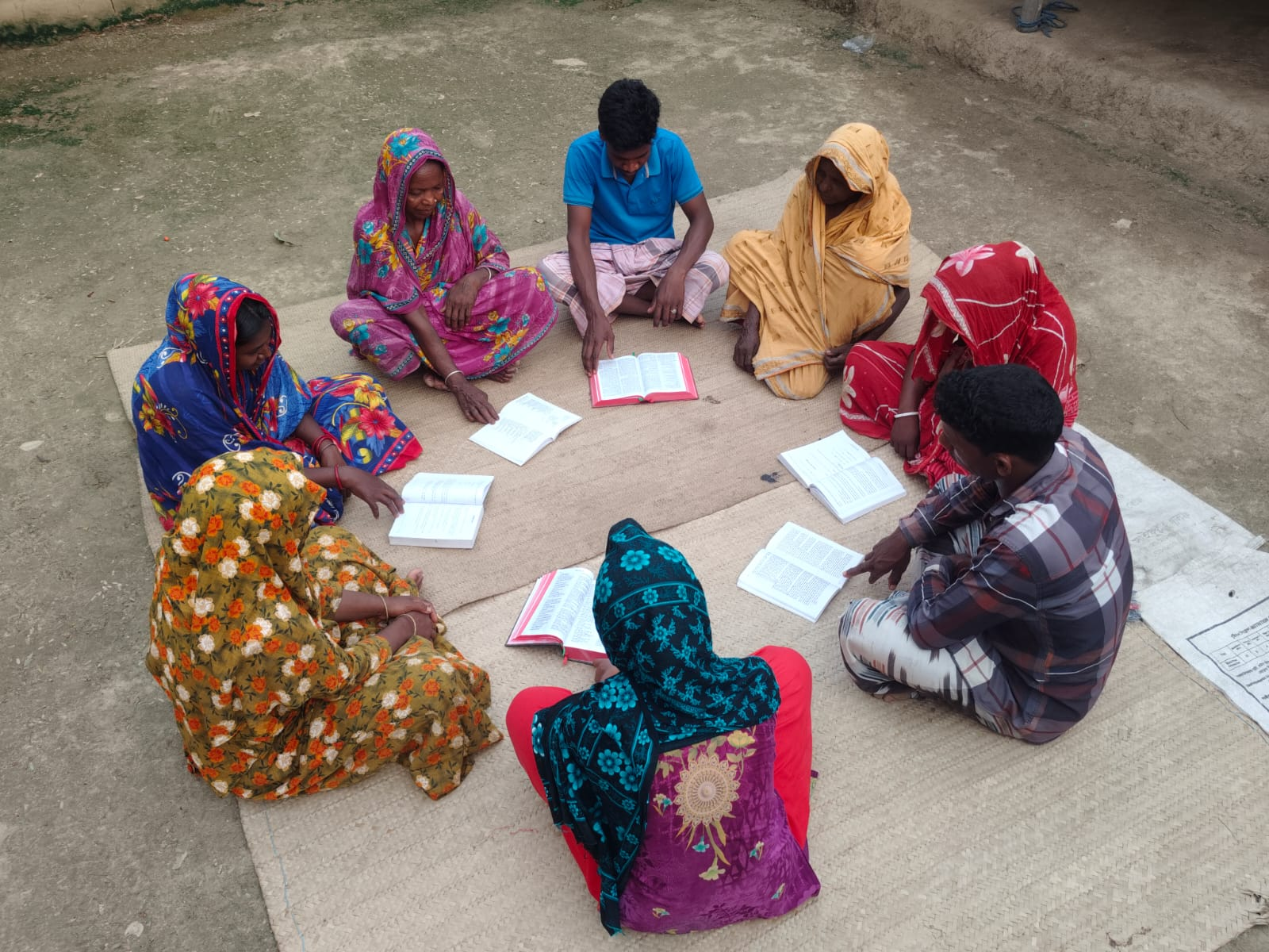 Bangla-language Bible study group seated outdoors in a circle with open Bibles